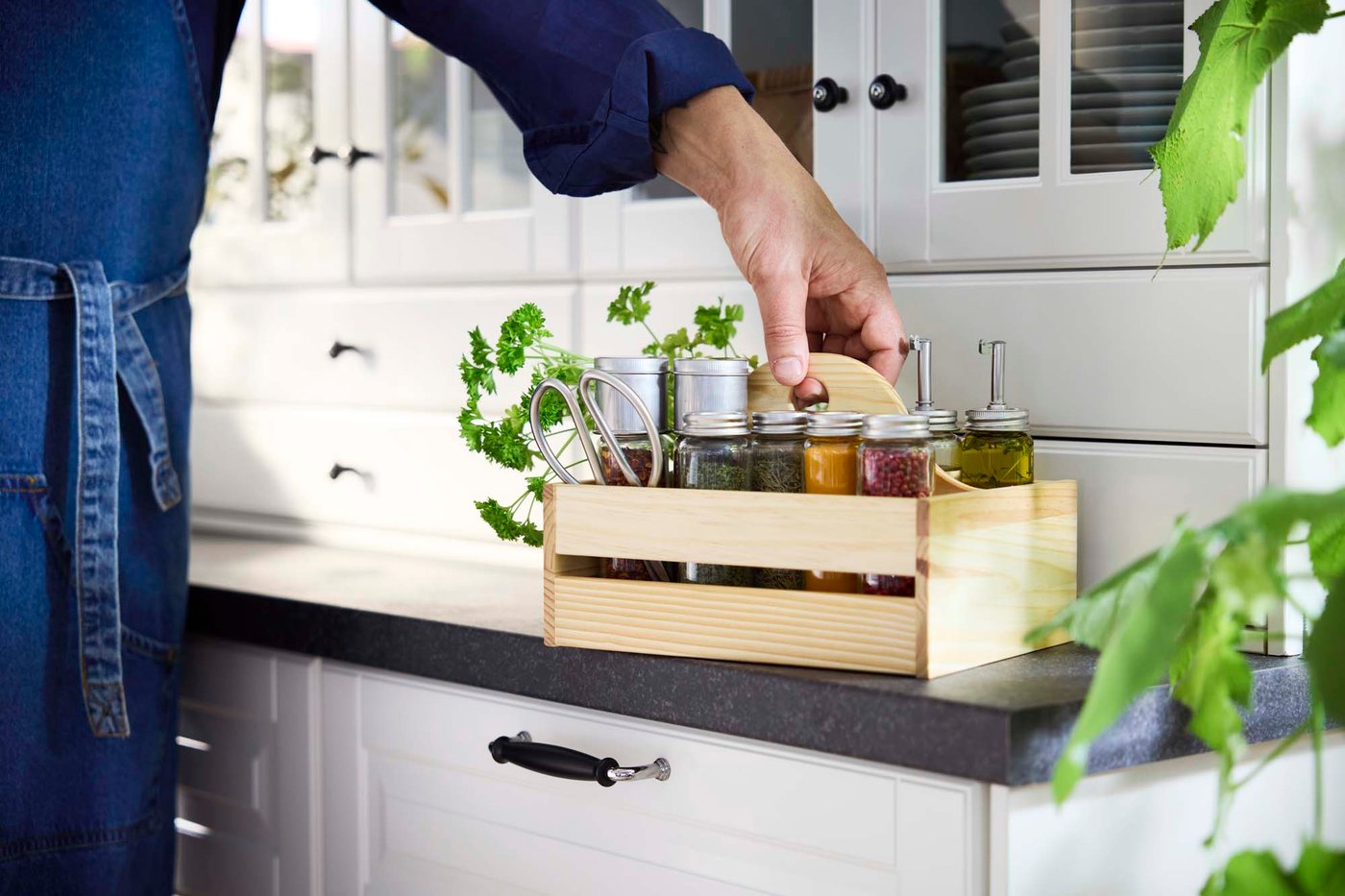 A person holds a light wood crate filled with herbs and spices in a white and dark grey kitchen. in