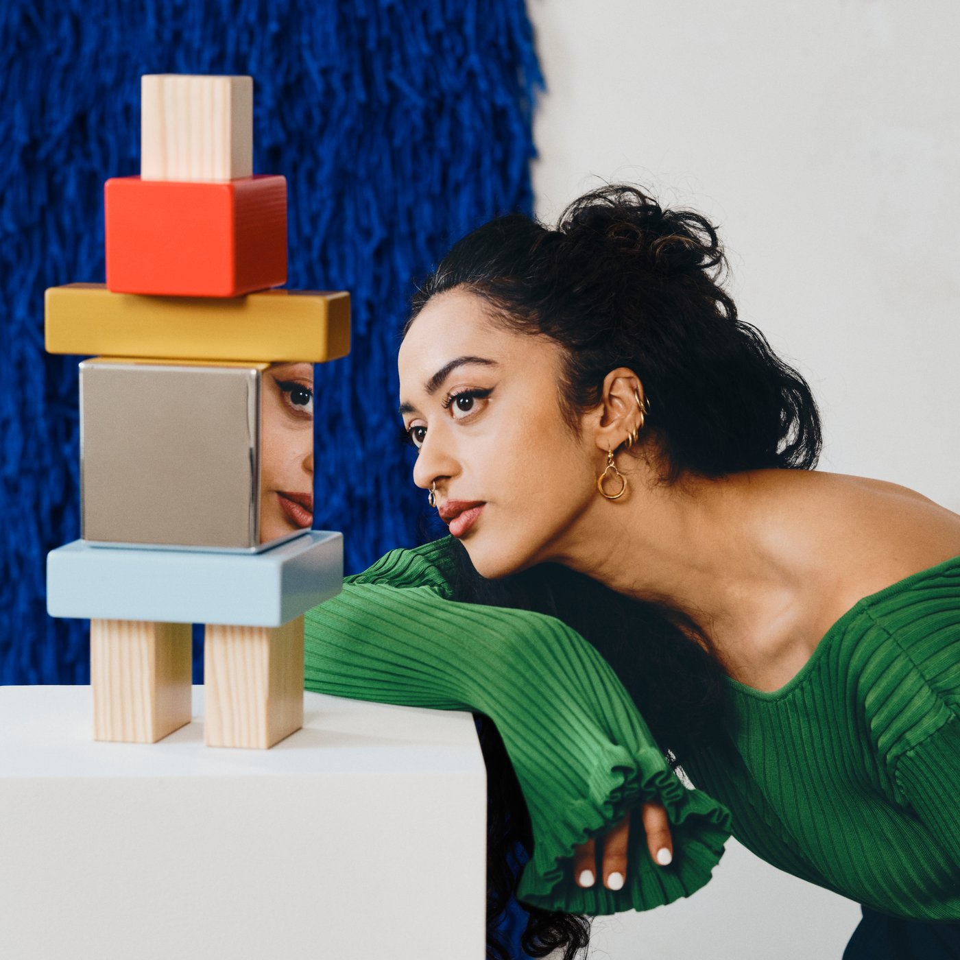 A woman in a green top leans beside a colorful geometric sculpture displayed on a white plinth in a contemporary art gallery.