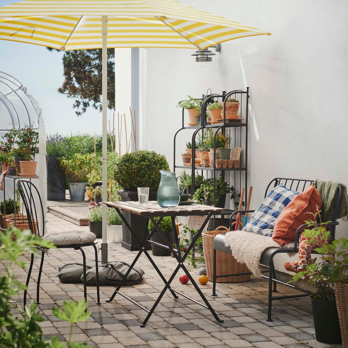 An outdoor dining setting with tables and chairs under a parasol in front of outdoor shelves and a greenhouse full of plants.