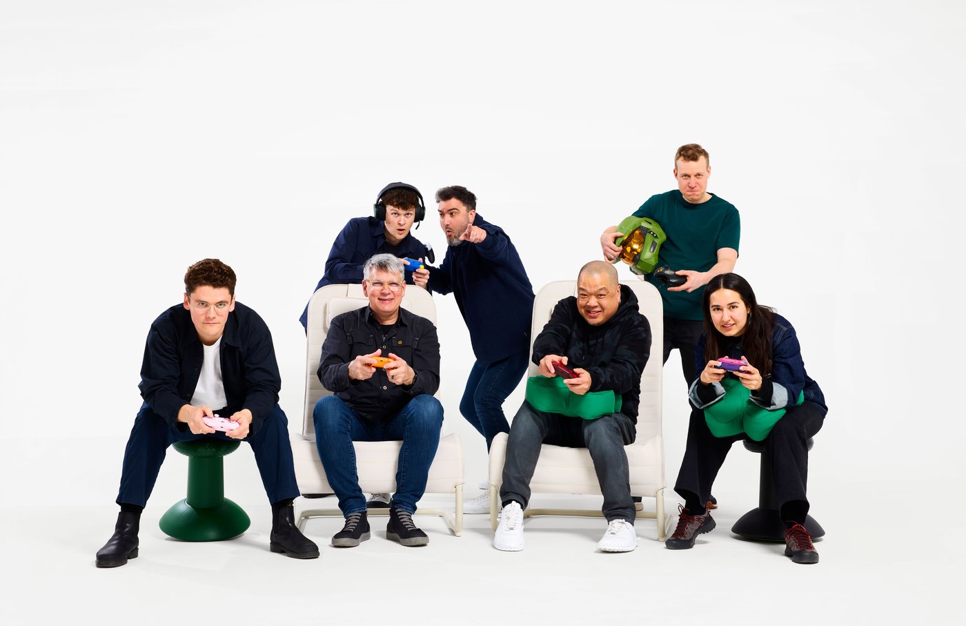 A group portrait of eight designers seated and standing around gaming-inspired furniture, holding controllers and gaming accessories against a white studio background.