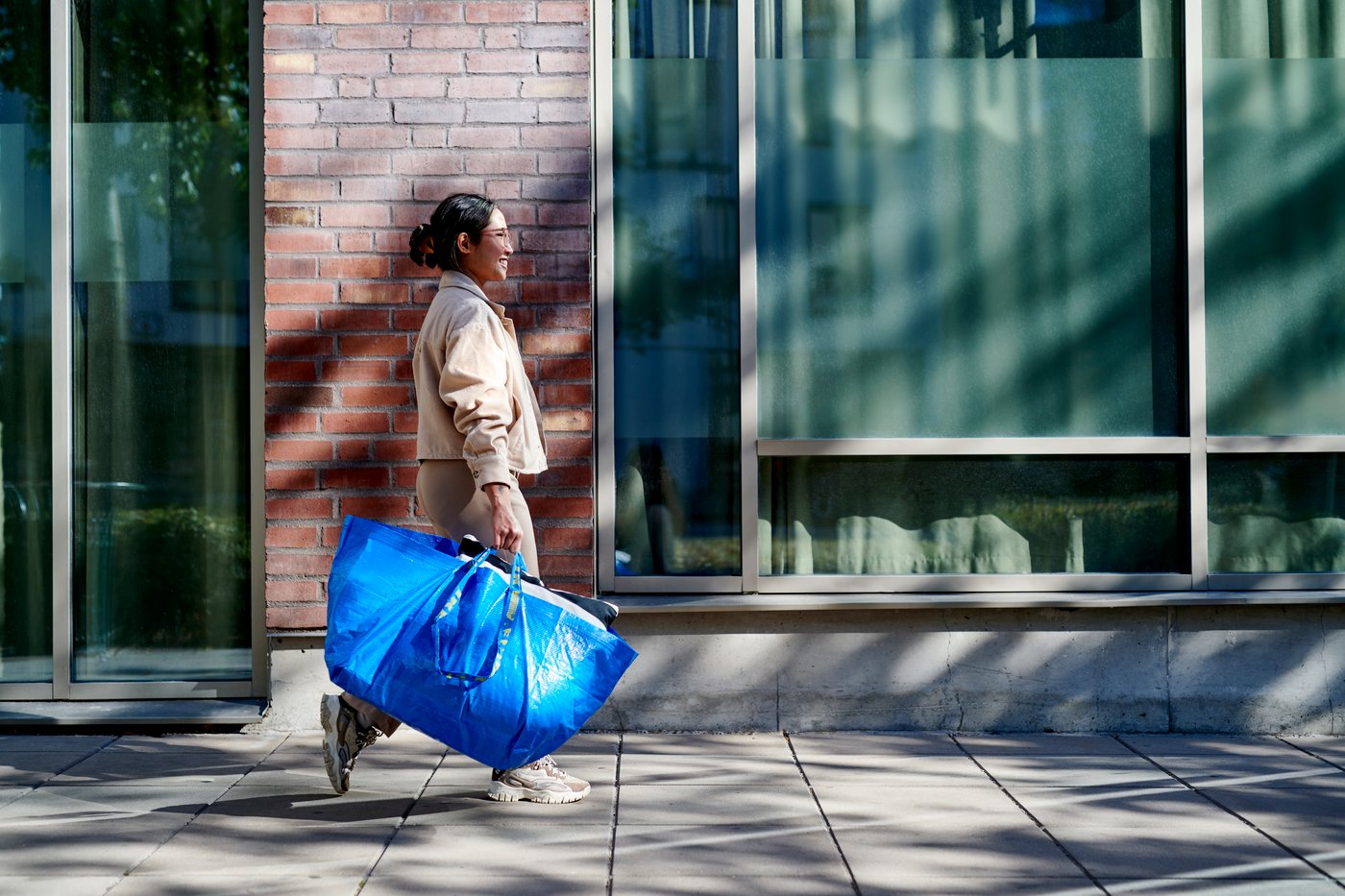 A smiling person walks on a sidewalk carrying a blue FRAKTA bag.
