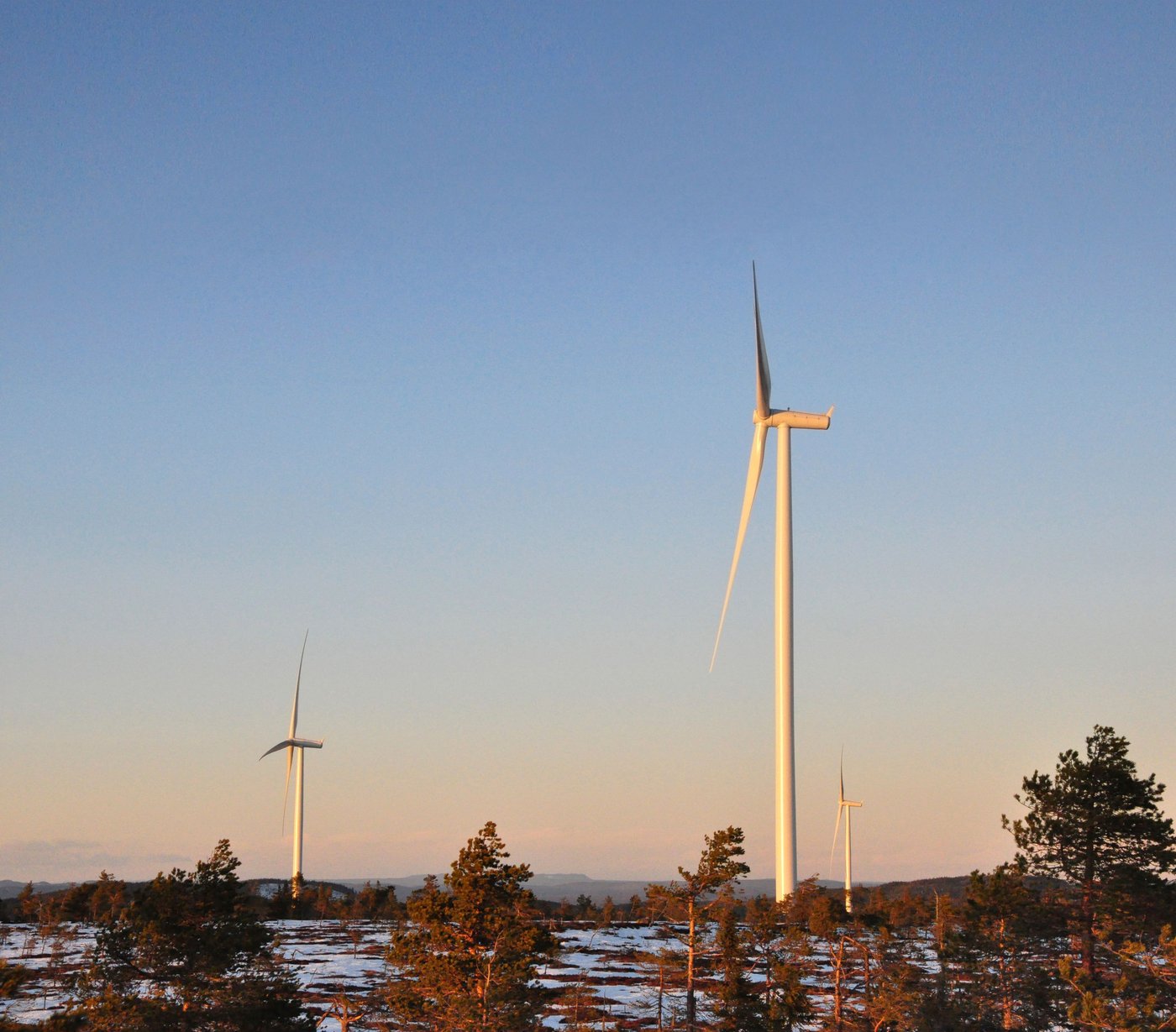 Three windmills with a blue sky and some trees in the foreground. 