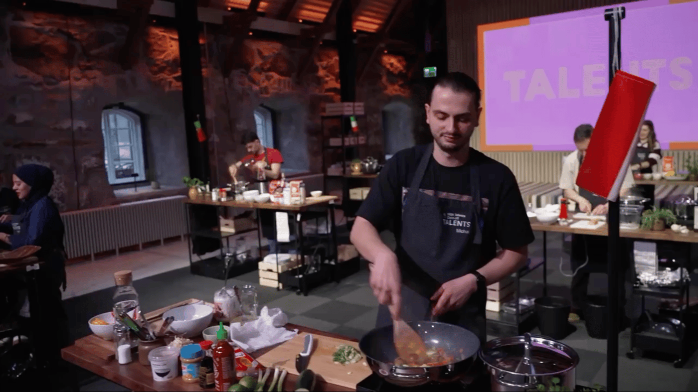 A person cooking in a stylish kitchen setting, with a wooden interior and brick walls. A screen in the background displays "TALENTS" in bold letters, while various kitchen tools and ingredients surround the workspace.