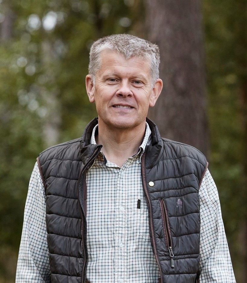 Portrait of Ulf Johansson who wears a checked shirt and a sleeveless puffer jacket, with trees visible in the background.