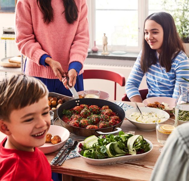 A family with two children around a dining table, having meatballs, mashed potatoes and a salad while talking and smiling.