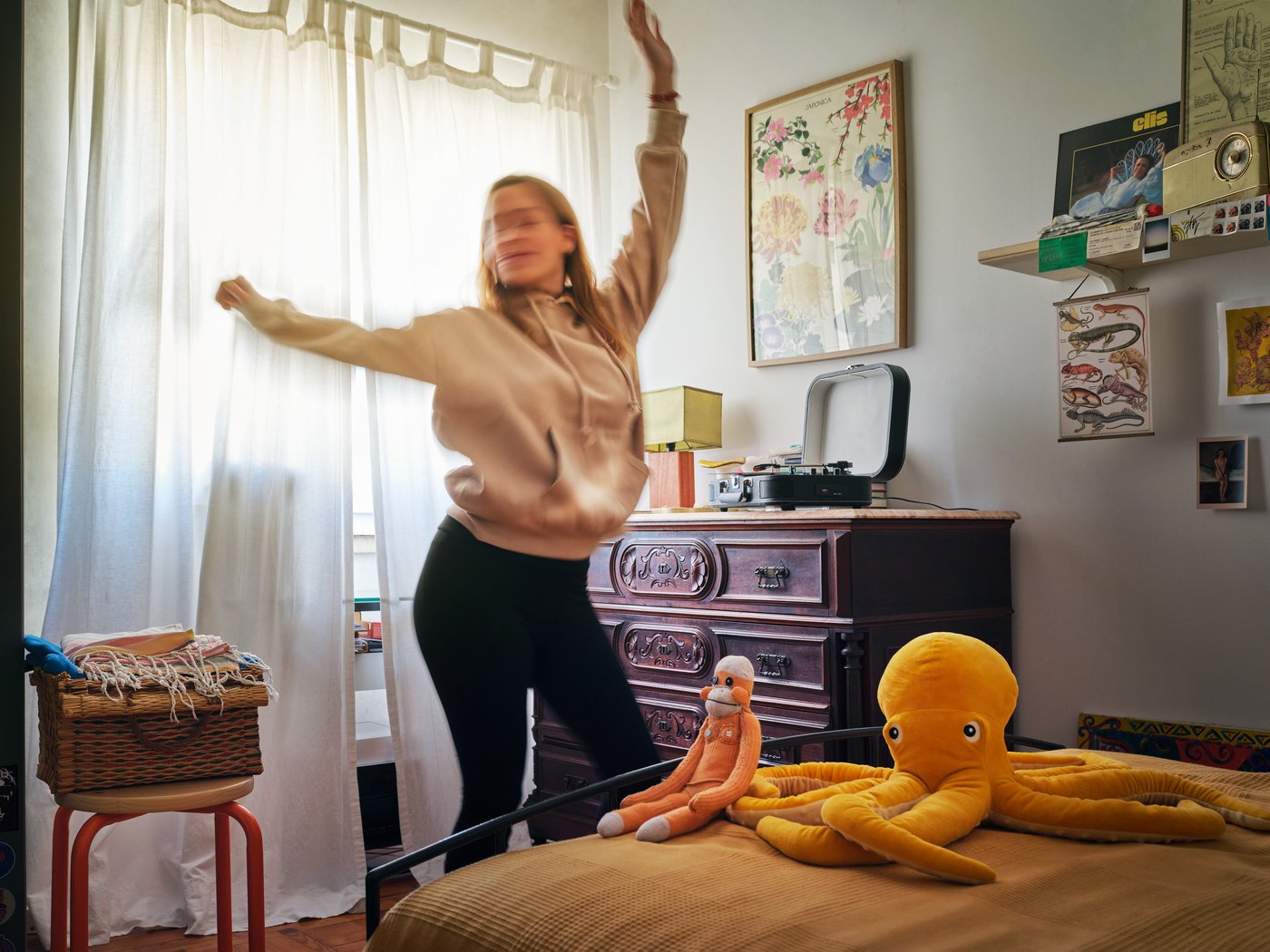A person dances in their bedroom. There are stuffed toys on the bed and pictures on the wall.