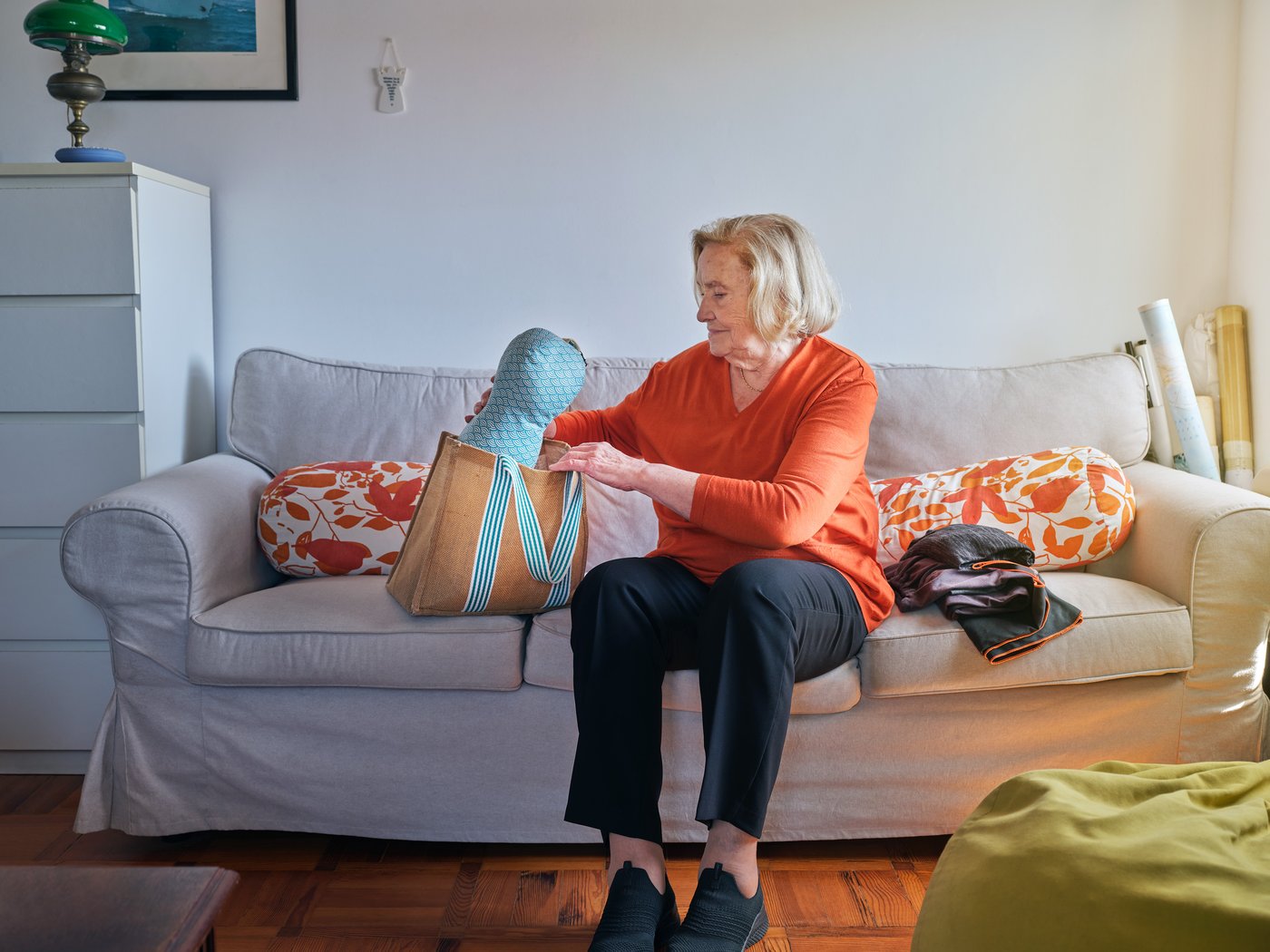 A person sits on a sofa in a living room. She is putting things into a fabric shopping bag.
