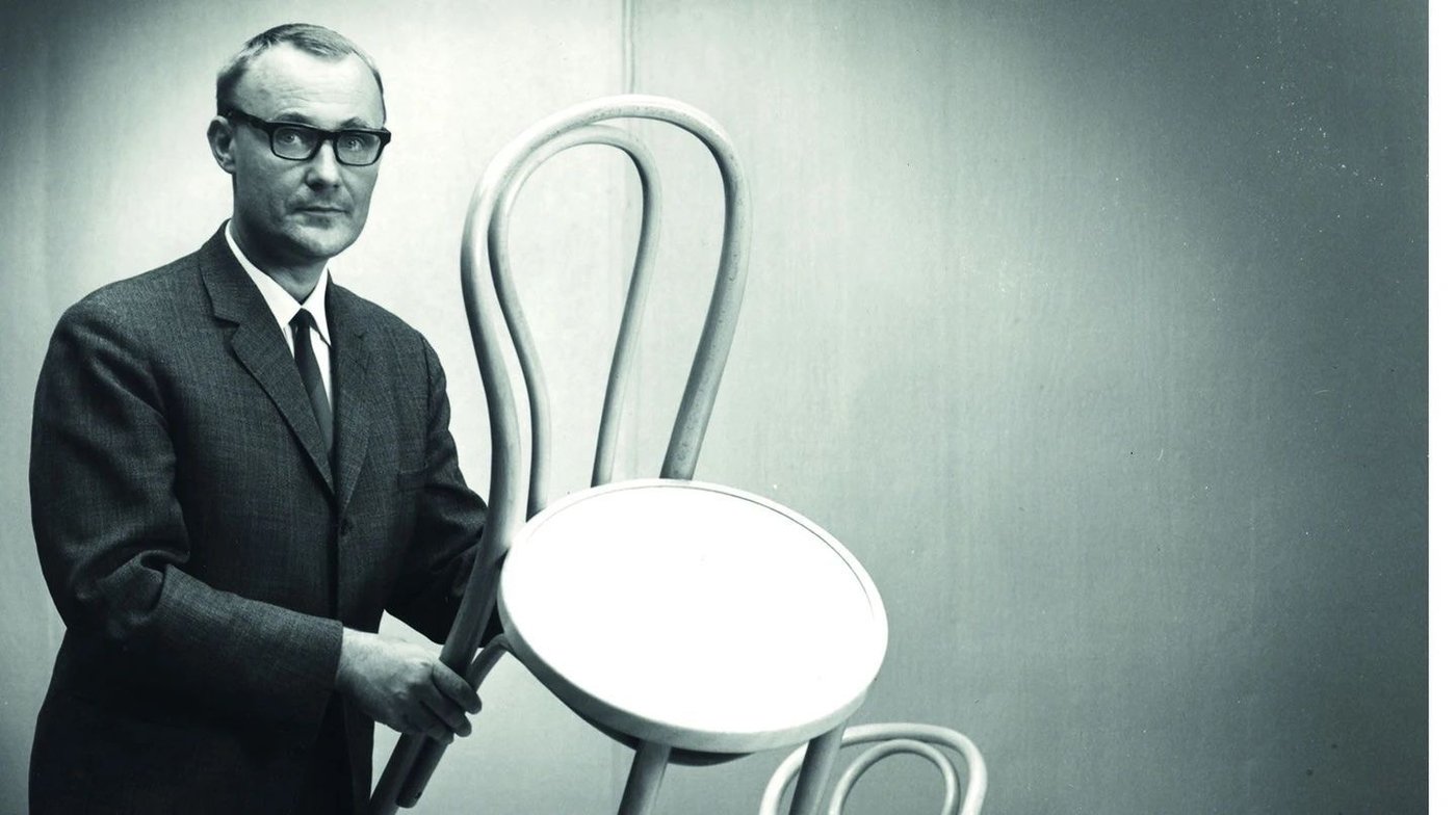Ingvar Kamprad in a suit and glasses, holding up a bentwood dining chair.