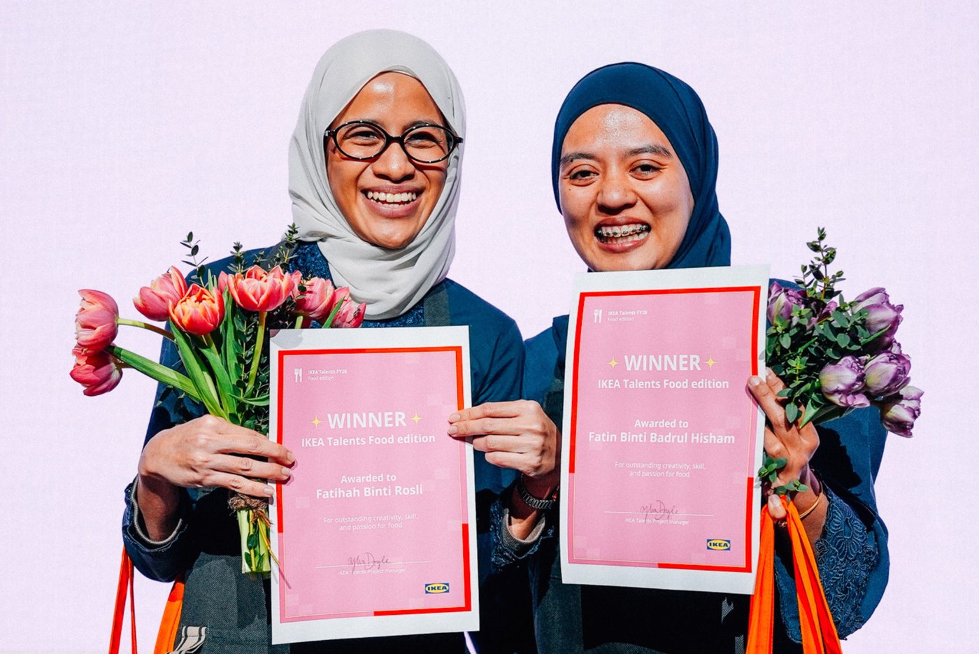 Two people smile at the camera as they hold their winner's certificate and a bunch of flowers.