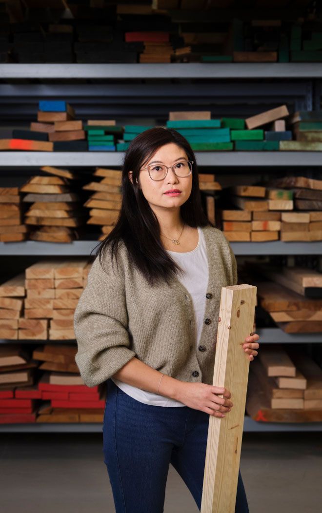 A woman stands in front of shelves stacked with wood. She is holding a piece of wood.