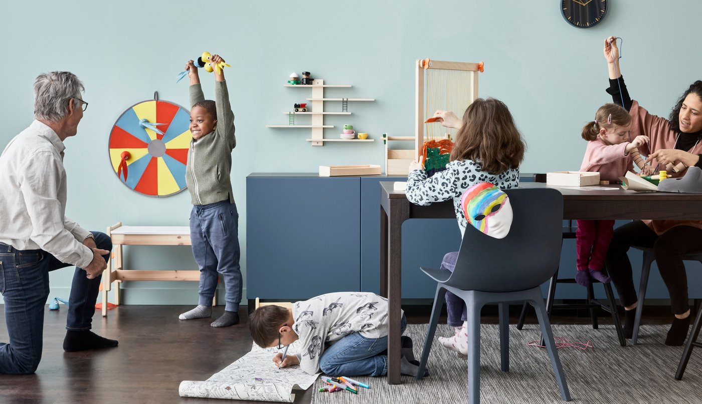 Two adults and two kids playing and drawing in a living room with a light blue wall and a blue cabinets.