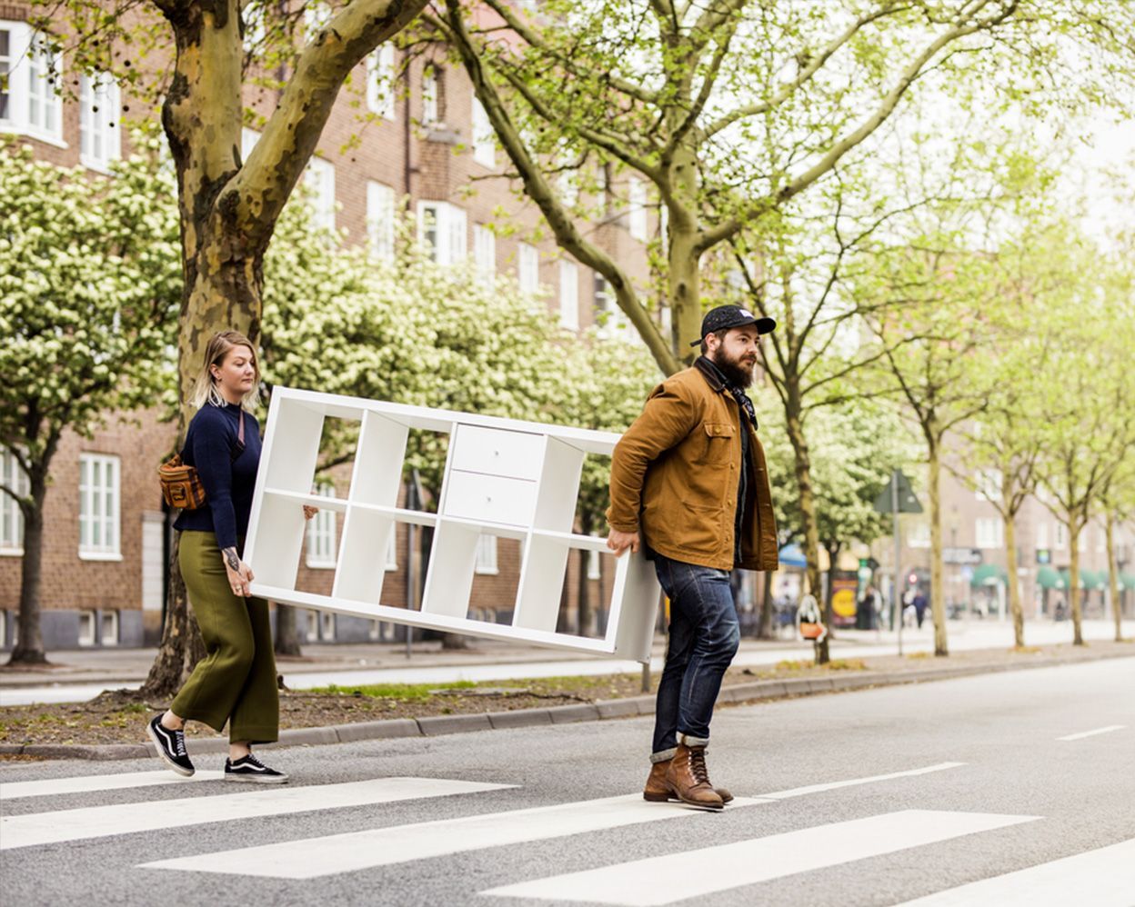 Two people carry a white shelving unit across a street.