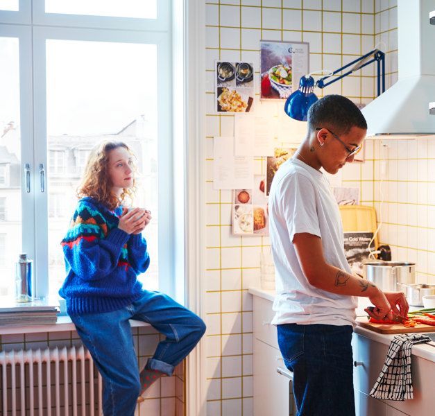 Two young people in a white kitchen with yellow accents, one prepares food and the other sits by the window, holding a cup.