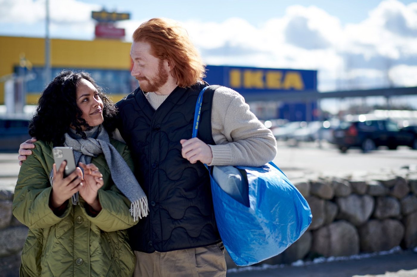 A couple are walking outside an IKEA store. They are smiling at each other. The man is carry a blue IKEA FRAKTA bag.