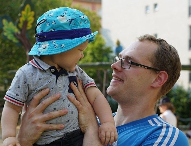 A man wearing glasses and a blue shirt holds and smiles at a baby wearing a blue bucket hat.