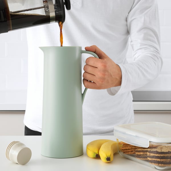 Person pours coffee into green insulated flask with partial lid. Table has bananas and cookies.