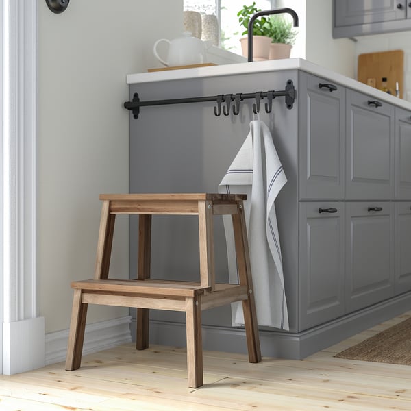 Wooden BEKVÄM step stool beside grey kitchen cabinets. Natural wood, small size.