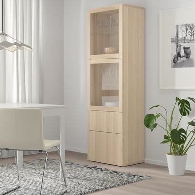 Light wooden BESTÅ cabinet with glass doors. Tall, narrow, fits well in corner. Stores dishes and glasses. Near white chair and plant.