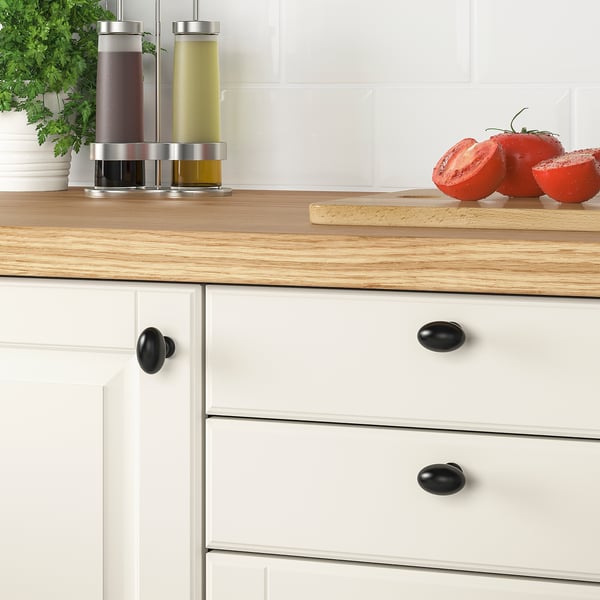 Kitchen counter with wooden top, white drawers, black knobs, bottles, tomatoes, and herbs.