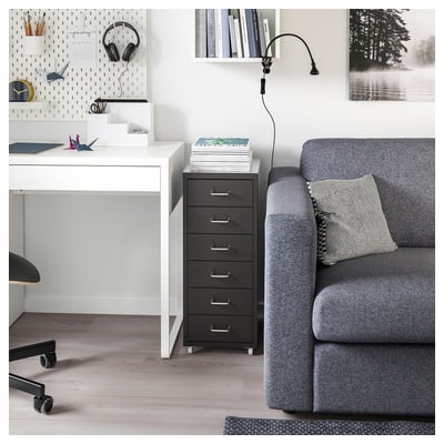 A HELMER black metal filing cabinet with five drawers on wheels beside a white desk and a grey armchair with a throw pillow.