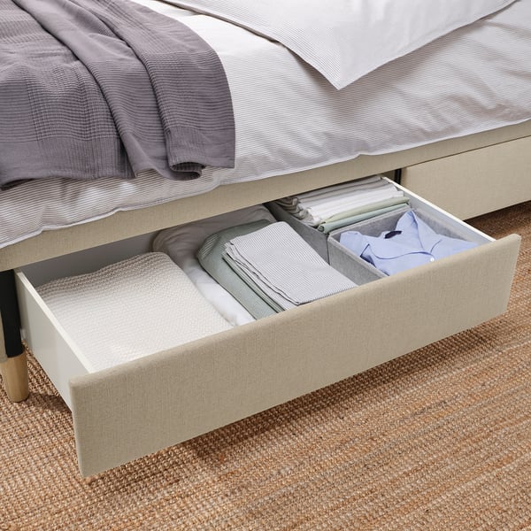 Bed frame with open drawer, neatly folded linens, soft-close feature, beige upholstery, wooden legs visible.