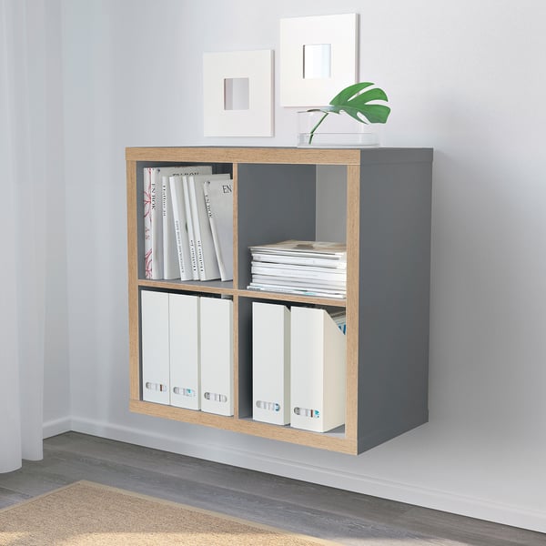 Gray KALLAX shelf with books, binders, plant, and empty frames in minimalist room.