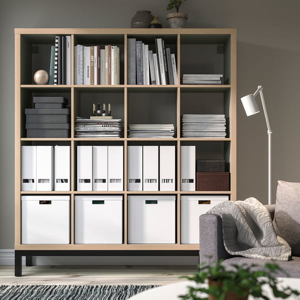 KALLAX shelving unit with books & decor. Tall, wooden, & spacious, divides room. Holds various items neatly. Large, rectangular shape.