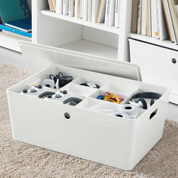 White organiser box with multiple compartments holding game controllers and cables, placed on rug near bookshelf.