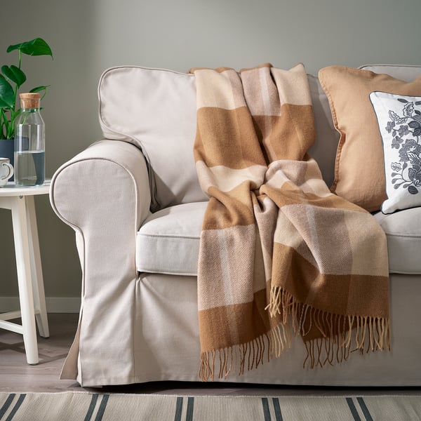 Beige sofa with brown-and-beige chequered wool throw and pillows, white side table, green plant.