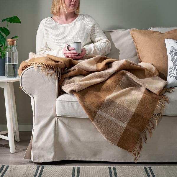 Person sitting on sofa with warm, light brown MYRULL wool throw blanket, holding a mug, enjoying cosy relaxation.