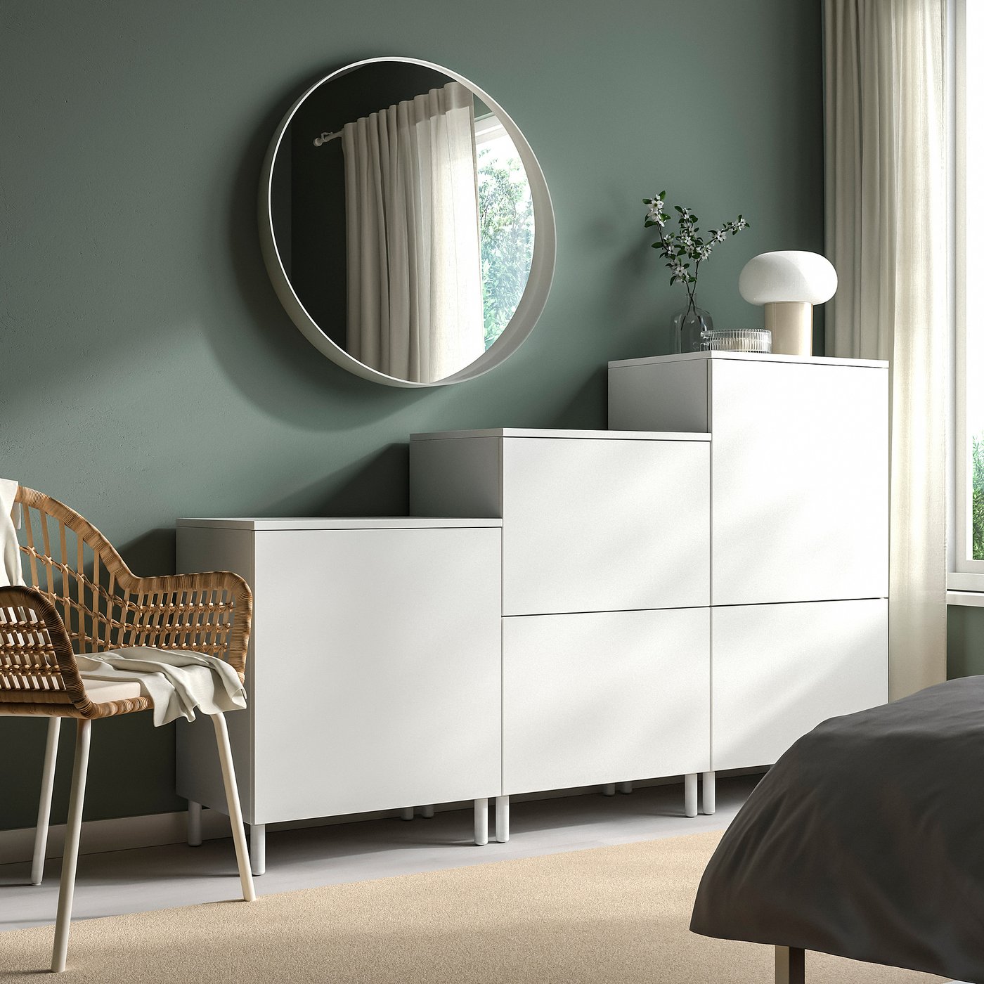 Minimalist bedroom with PLATSA white storage units, round mirror, wicker chair, and grey bed against sage green wall.
