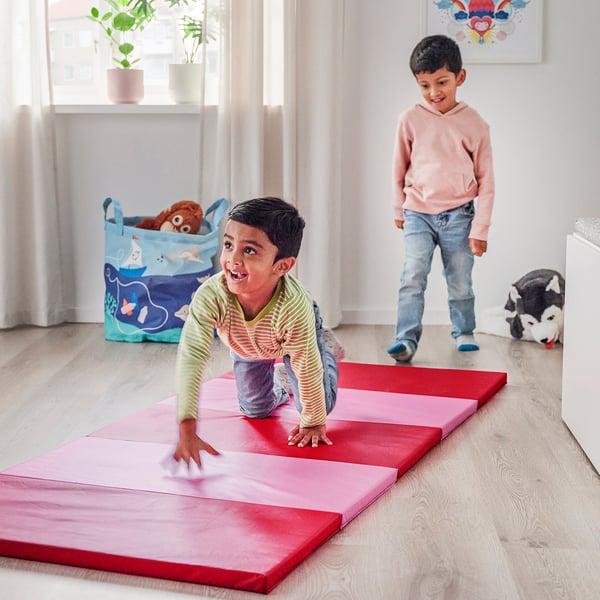 Kids playing indoors on colourful mat; one does somersaults, other jumps.