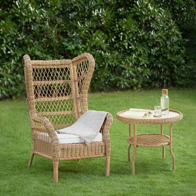 Wicker chair & side table on lawn, rattan, cushion, blanket, glassware, book, green bushes.