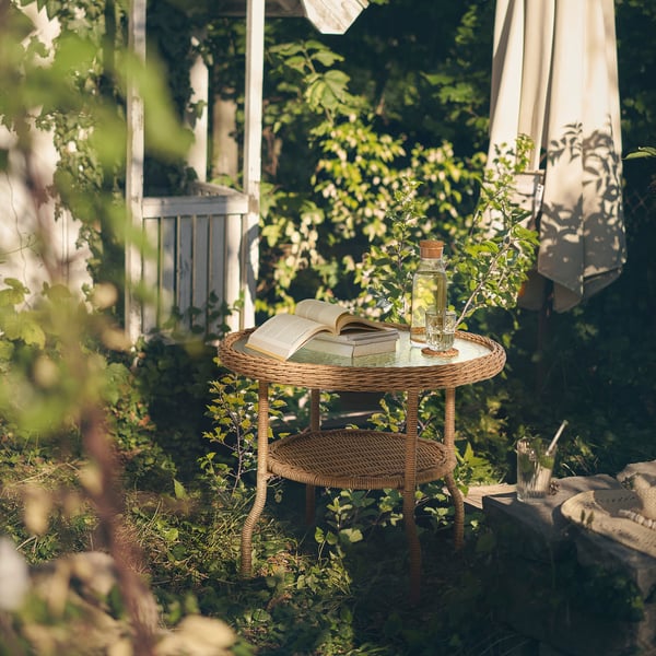 Round, wicker RISHOLMEN coffee table with glass top, holding book & drink, set in green garden.