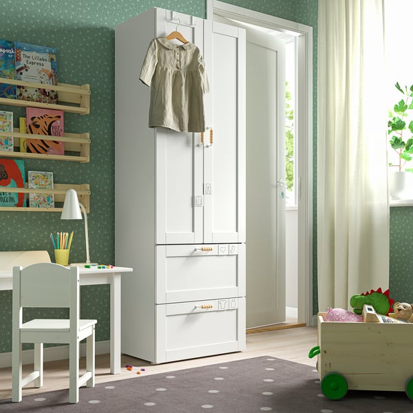 A kids room with a white SMÅSTAD wardrobe, a small desk, and toys. A dress hangs on the wardrobe, and books line the wall.