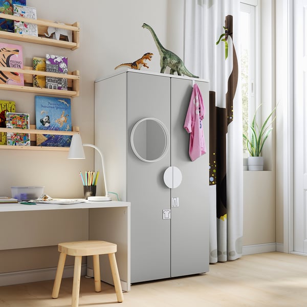 Kids room with light grey SMÅSTAD wardrobe, dinosaur toys, wooden stool, bookshelf with colourful books.
