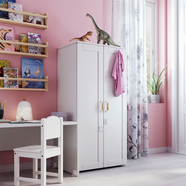 Childs room with white SMÅSTAD wardrobe, pink walls, wooden shelves filled with colourful books, dinosaur toys on top, white desk, chair, and lamp.