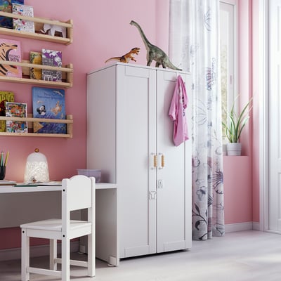 Childs room with white SMÅSTAD wardrobe, pink walls, wooden shelves filled with colourful books, dinosaur toys on top, white desk, chair, and lamp.