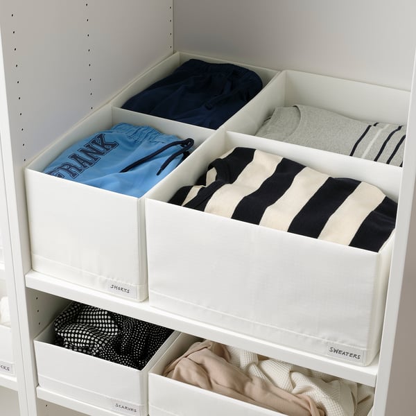 White storage shelves with labelled boxes holding neatly folded clothes, showcasing organised storage for different types of garments.