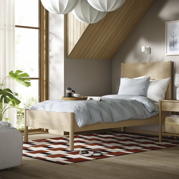 Modern wooden bed, white pillows, gray sheets. Nightstand with lamp & plant. Colorful rug, big window, trendy.