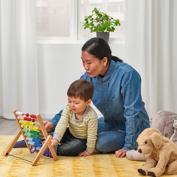 A person and child play with a colourful, foldable abacus on a yellow rug, surrounded by soft toys.