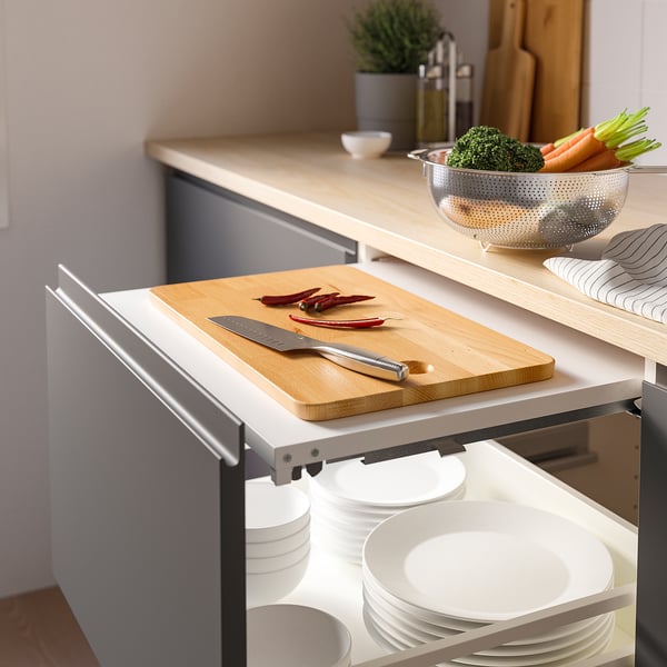 Kitchen counter with pull-out cutting board holding knife and veggies, next to plates and bowls in drawers.