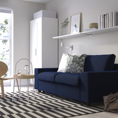 Modern living room with blue VIMLE sofa, white rug, wooden side table, and shelf with books and decorations.