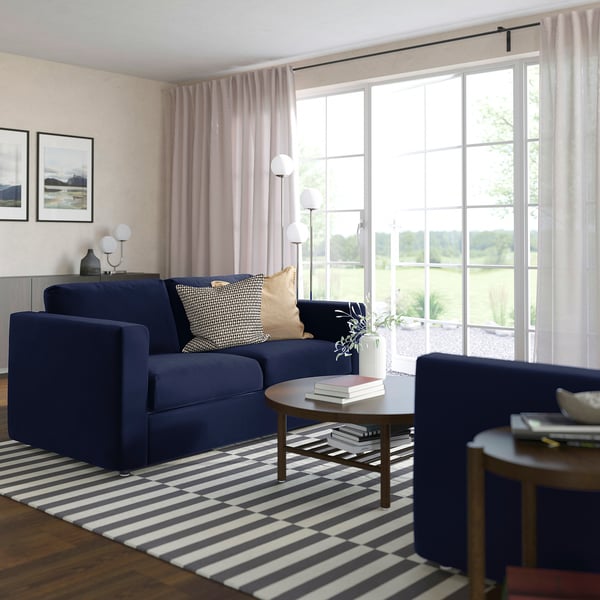 Blue VIMLE sofa, modern minimalist design, large windows, striped rug, wooden tables, cosy and spacious living room.