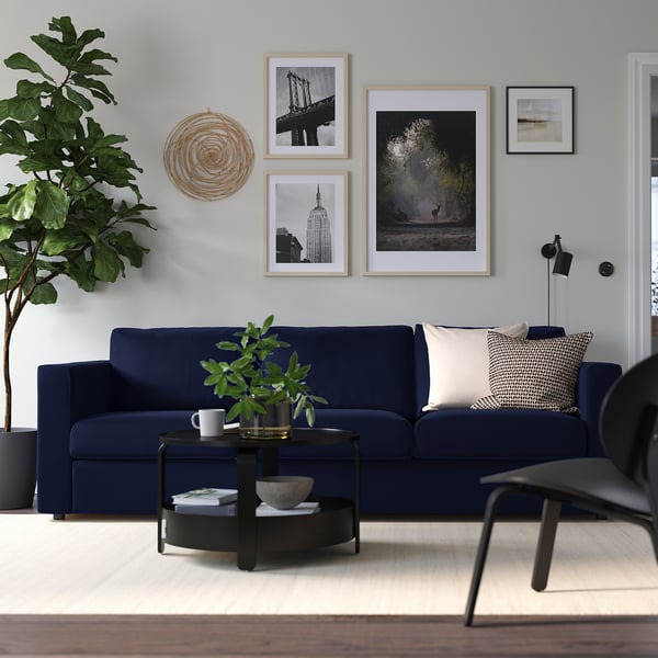 Cosy living room with navy VIMLE sofa, beige cushions, black coffee table, and minimalist art. Large houseplant adds greenery.