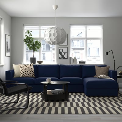 Modern living room with blue VIMLE sofa, rug, pendant light, coffee table, chair, plant, and bookcase. Natural light from window.