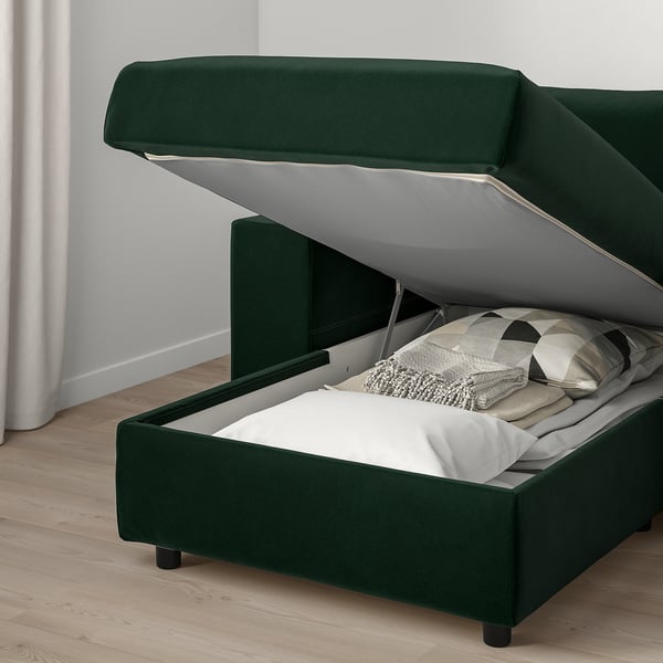 Green upholstered sofa with hinged section revealing storage compartment under seat, white bedding inside.