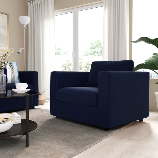 Modern living room with large navy blue sofa. Soft cushions, wooden floor, and natural light. Large windows with white curtains.