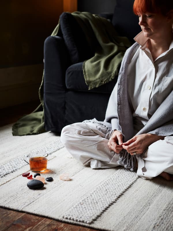 밝은 색상의 러그가 깔려 있고 앞쪽에 크리스털, 조약돌, 유리 머그잔이 놓여 있는 거실에 앉아 있는 여성.