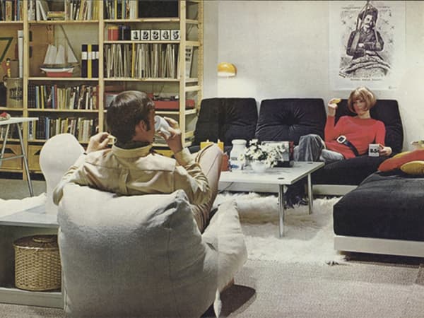 A 1980’s living room with two seated people, with several IVAR shelving units in the background.